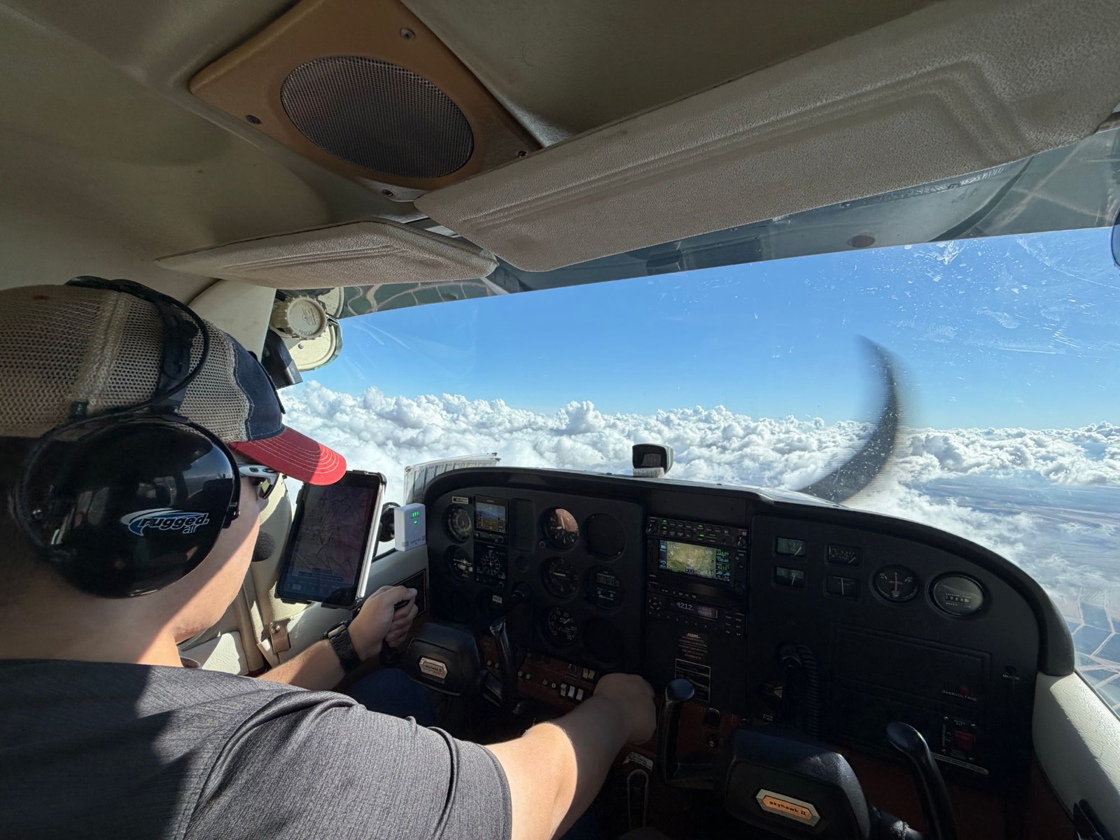 A pilot in a Cessna 172 cruising above a solid layer of white clouds under a blue sky, iPad chart mounted on the yoke